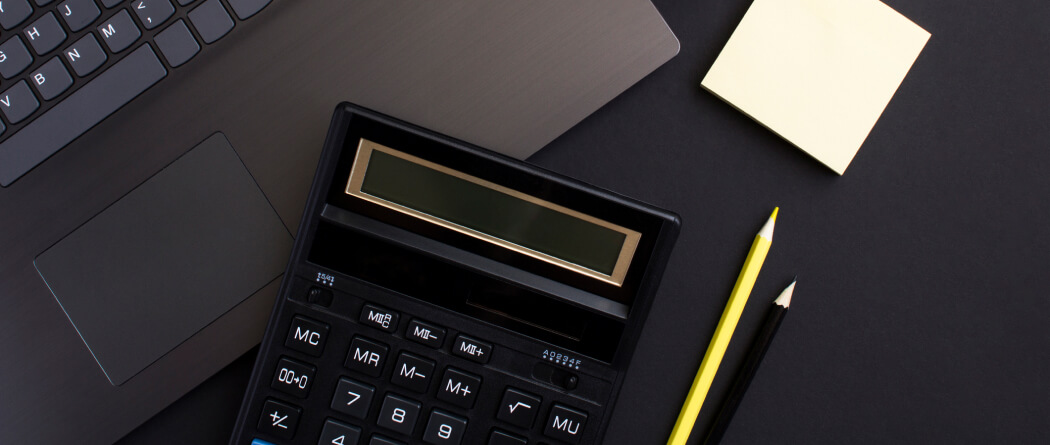 A top-down view of a calculator, laptop, sticky notes, and pencils on a black surface. 